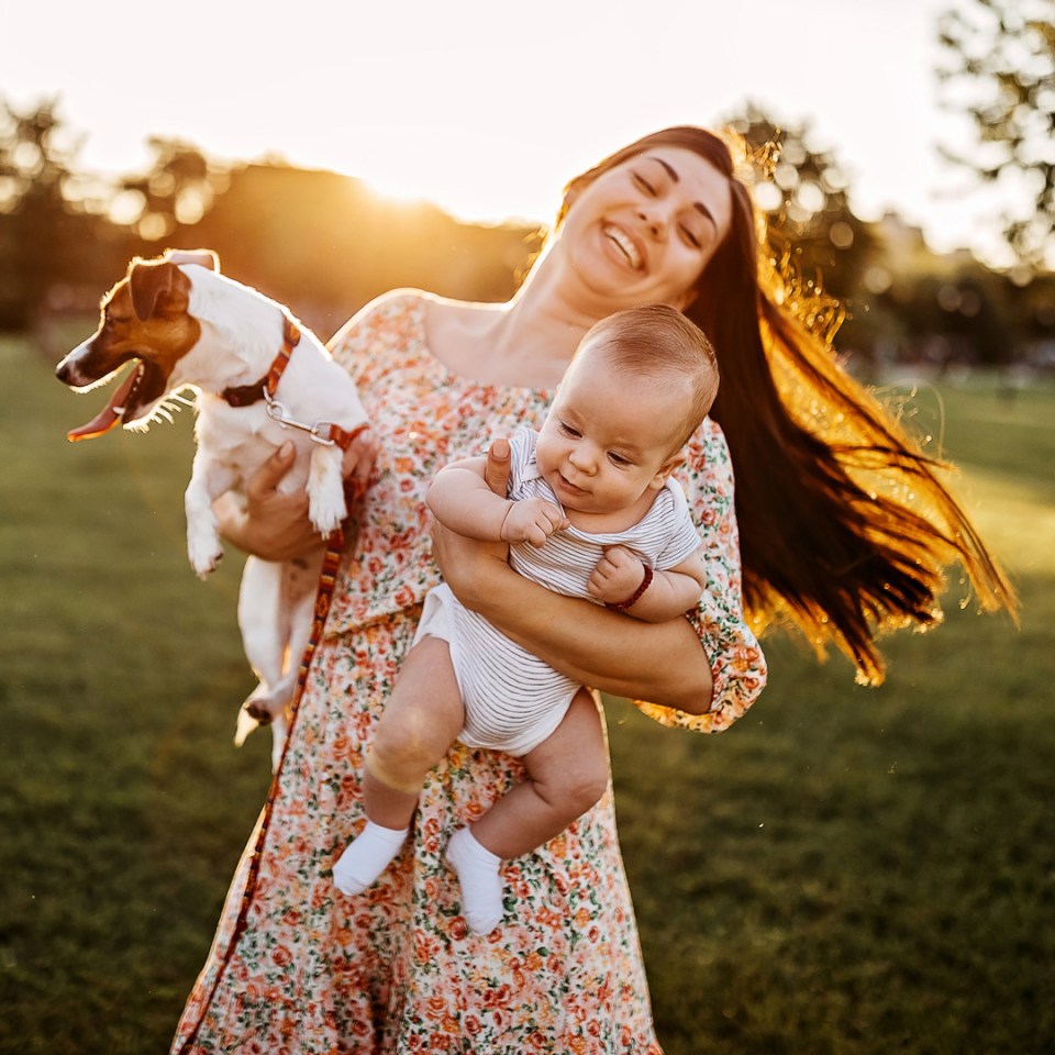 Une maman tenant son enfant et son chien dans un parc avec un joli coucher de soleil.